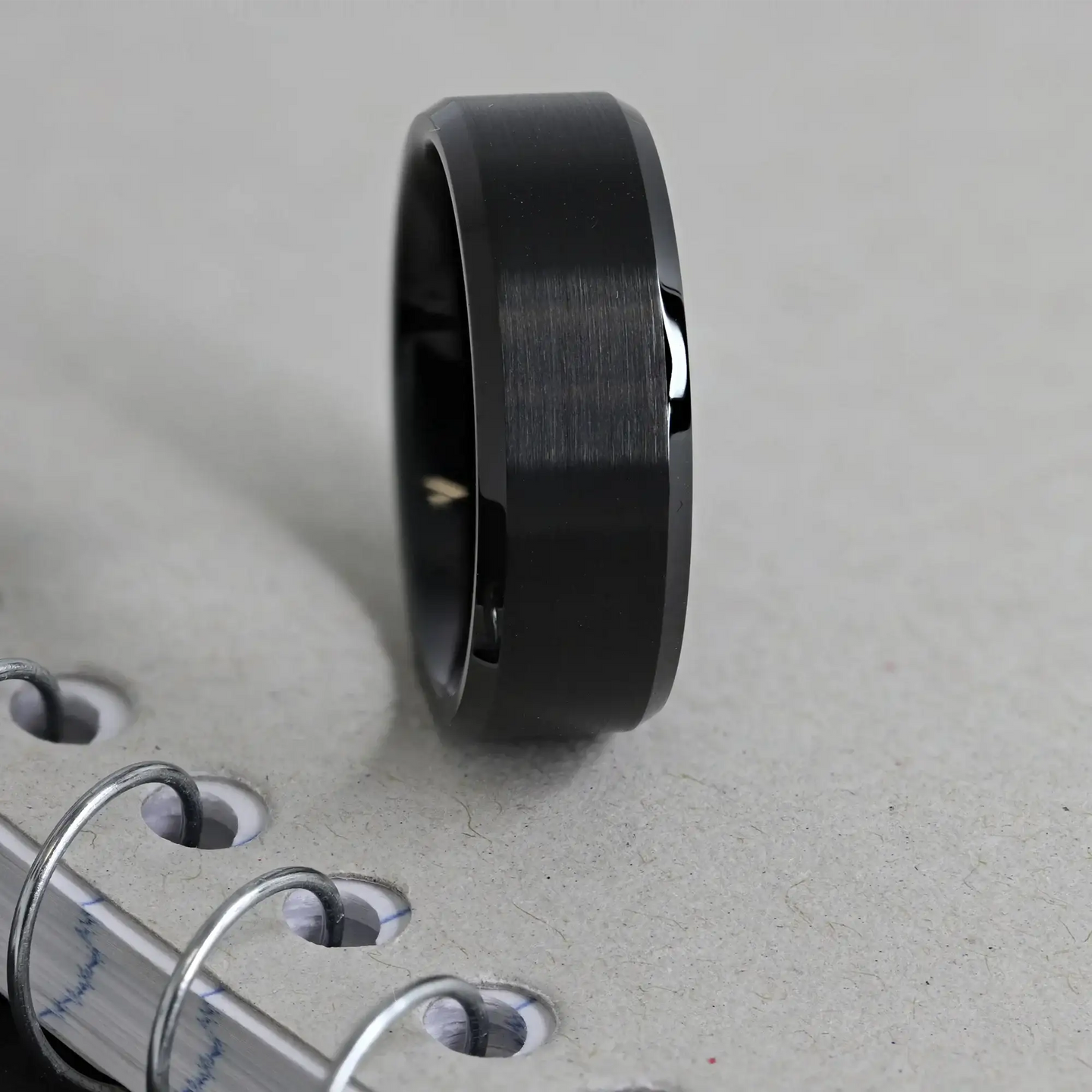 Lifestyle shot of the black brushed tungsten ring standing on a spiral notebook