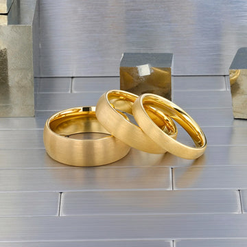 Three brushed gold-plated tungsten rings placed on a wooden desk with papers and a pen