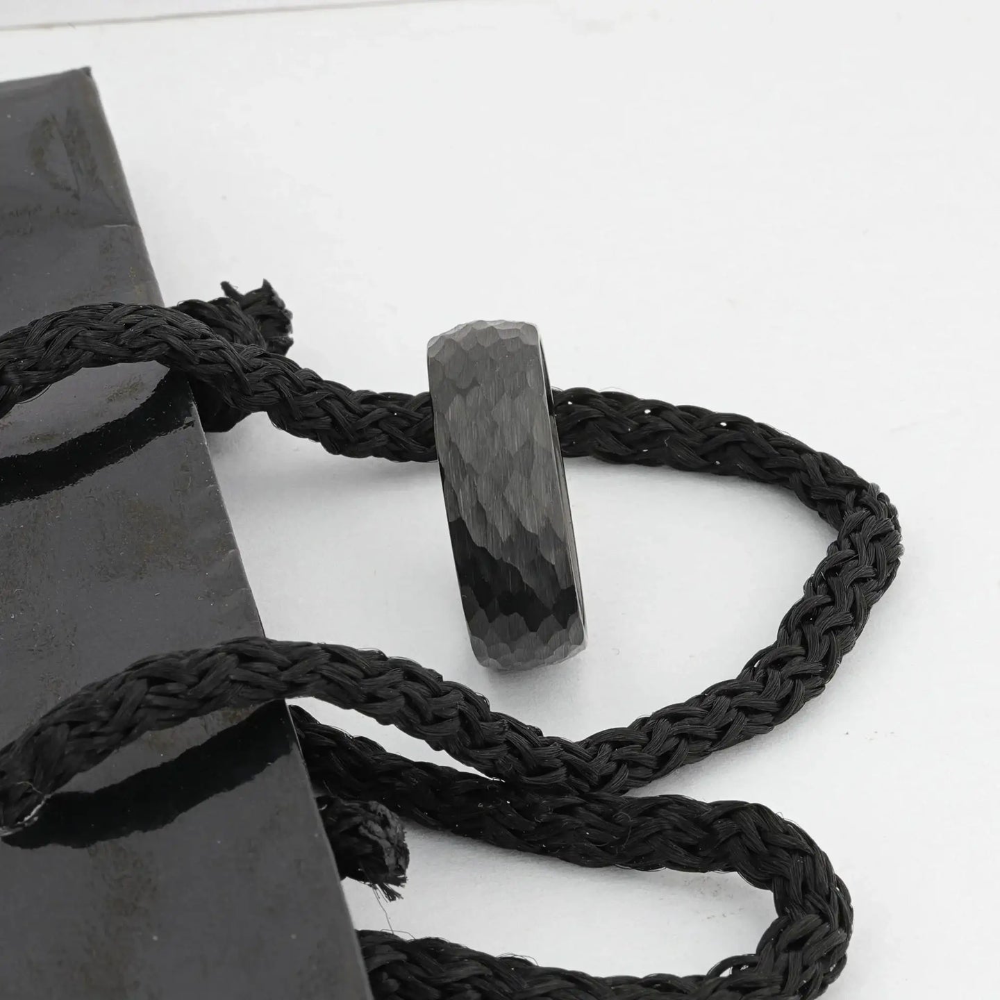 Close-up lifestyle image of the RENEGADE hammered black tungsten carbide band resting on braided cord