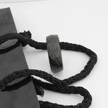 Close-up lifestyle image of the RENEGADE hammered black tungsten carbide band resting on braided cord