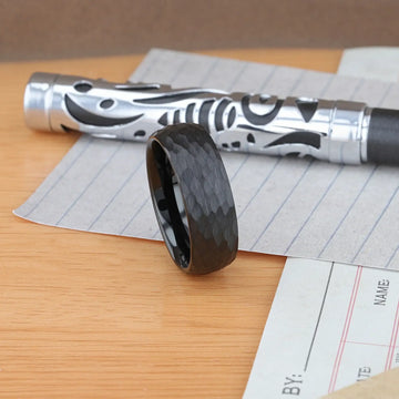 Desk scene showing the RENEGADE hammered black tungsten carbide wedding band beside a pen