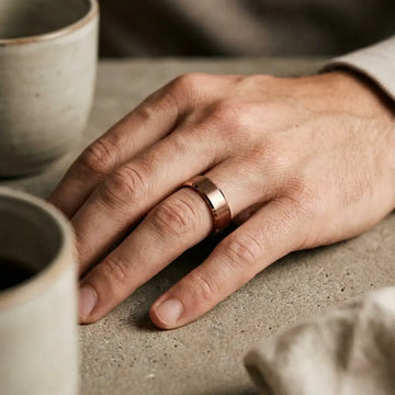Two-tone tungsten carbide ring with flat beveled edges and brushed center band worn on person's hand next to ceramic cup.