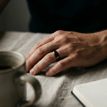 Dark brown domed wedding band on man's hand resting on fabric surface.