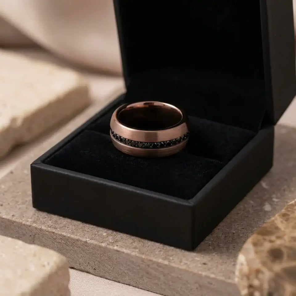 Tungsten carbide band with dark inlaid stripe displayed in black ring box on marble surface.