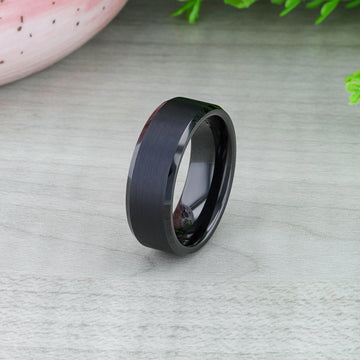 Black tungsten ring with brushed center displayed on a wood-textured surface with greenery