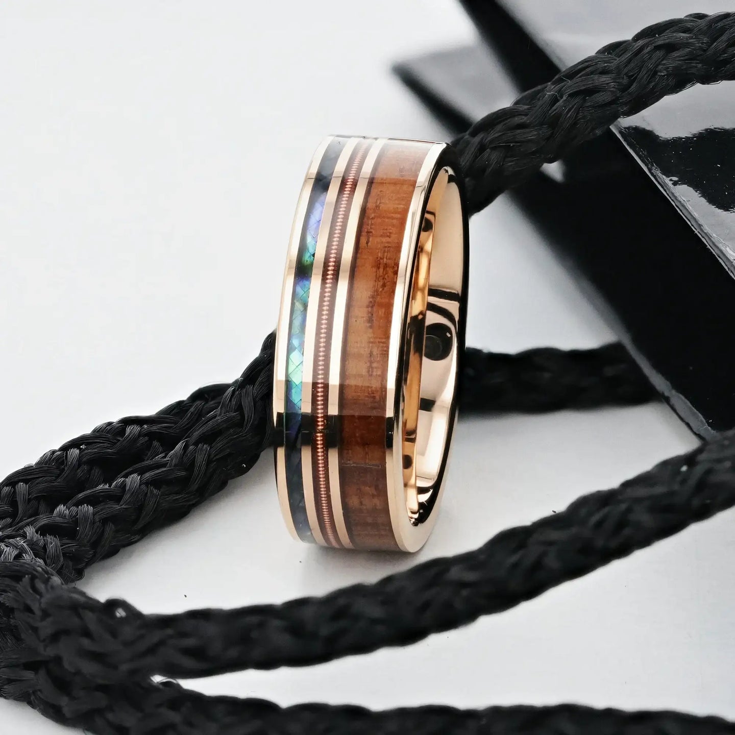 Lifestyle shot of the smoked rose gold tungsten ring with koa wood, abalone, and guitar‑string inlays resting on braided rope