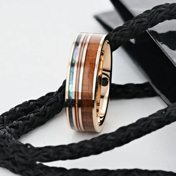 Lifestyle shot of the smoked rose gold tungsten ring with koa wood, abalone, and guitar‑string inlays resting on braided rope
